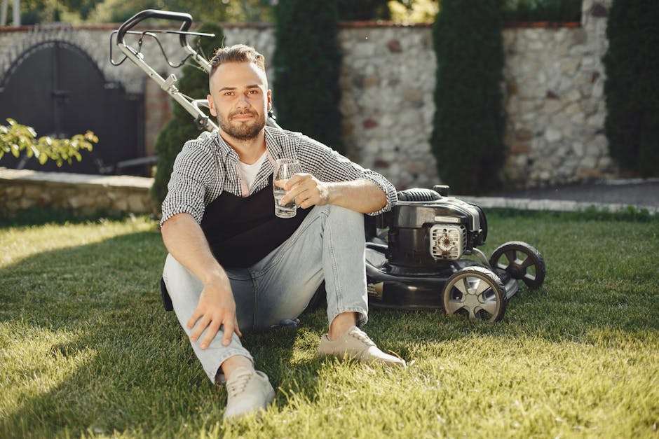 A young man with a beard and short hair, sitting on a well-maintained lawn in a residential garden during daylight hours, holding a glass of water and smiling at the camera. Behind him, a lawnmower wi
